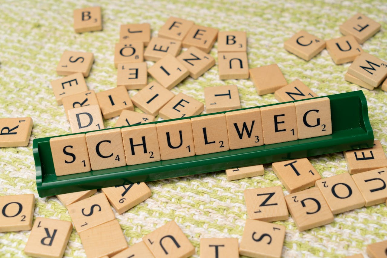 about-me-img Scrabble tiles spelling Schulweg on a rack, surrounded by scattered letters on a knitted background.